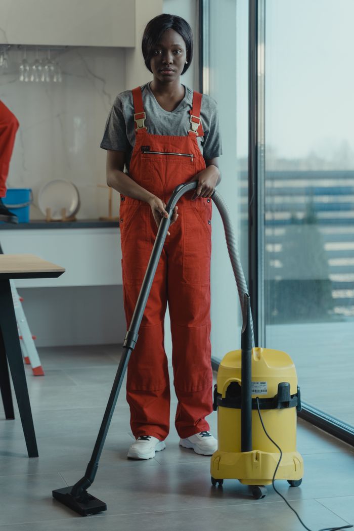 services-01 An adult woman in red coveralls operates a vacuum cleaner in a modern indoor setting, focusing on cleaning tasks.